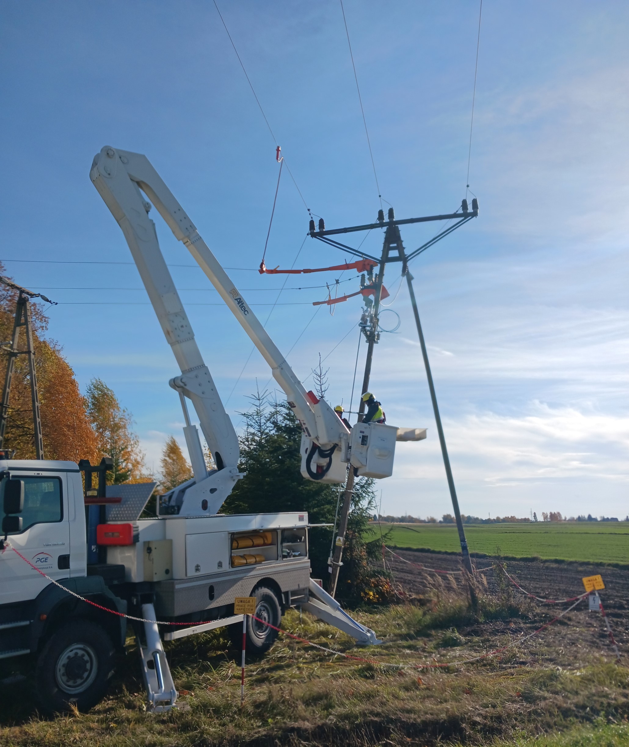 dzień BHP. Słup linii średniego napięcia, pod nim podnośnik z wysuniętym kosztem; w koszu dwóch elektromonterów
