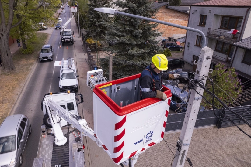 widok z g&oacute;ry - elektromonter w koszu, wysoko przy wierzchołku słupa energetycznego, naprawia linię zasilającą. W dole ulica, jadące auta