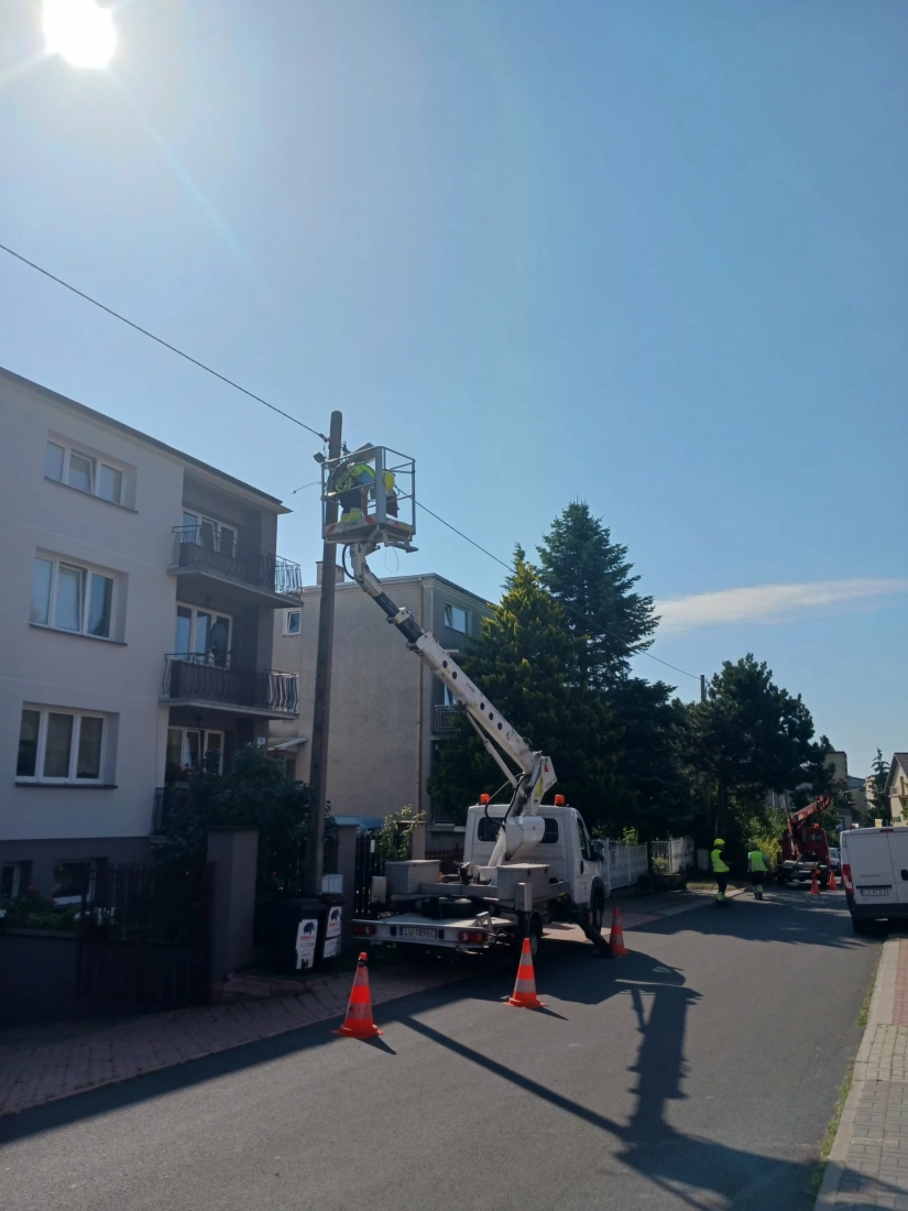 ulica, domy, chodnik. Na chodniku stoi rozłożony podnośnik z koszem. W koszu elektromonter naprawia linię zasilającą na słupie
